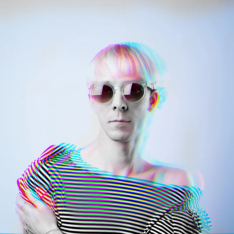 Porträt von Coco Schwarz mit runder Sonnenbrille und gestreiftem Shirt, das Bild in schillernde Regenbogenfarben verfremdet.